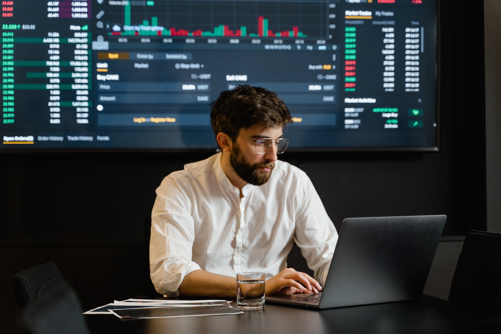 Dark-haired man in white shire and a pair of glasses working on a laptop in front of a large monitor showing various data.